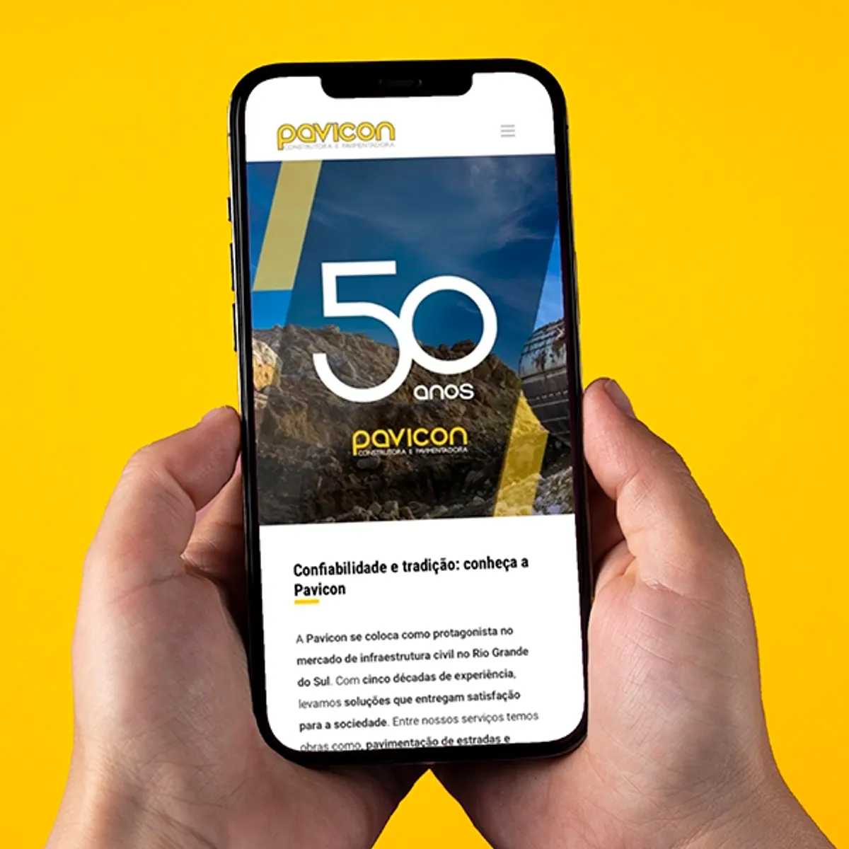 Mãos segurando um smartphone que exibe o site da Pavicon desenvolvido pela W3COM. A página mostra o aniversário de 50 anos da empresa e seu foco em confiabilidade e tradição na infraestrutura civil. Este exemplo mostra como a W3COM entrega soluções estratégicas e eficazes para o mercado B2B.